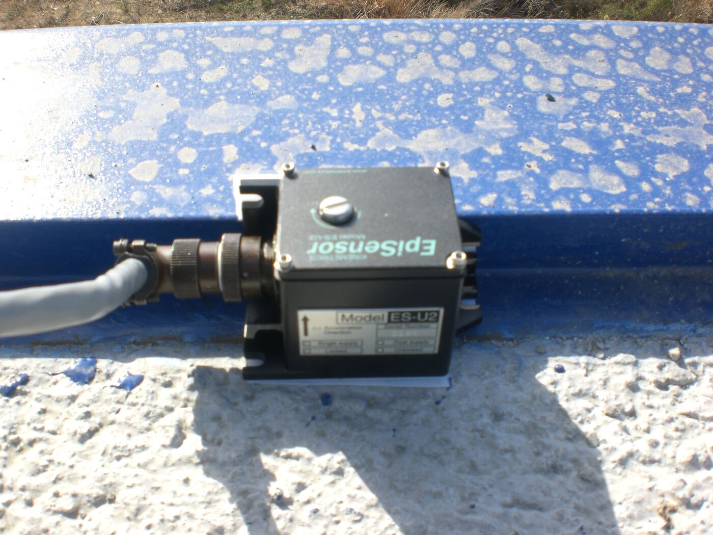 Acelerómetro negro con conector, fijado sobre una barandilla metálica azul
