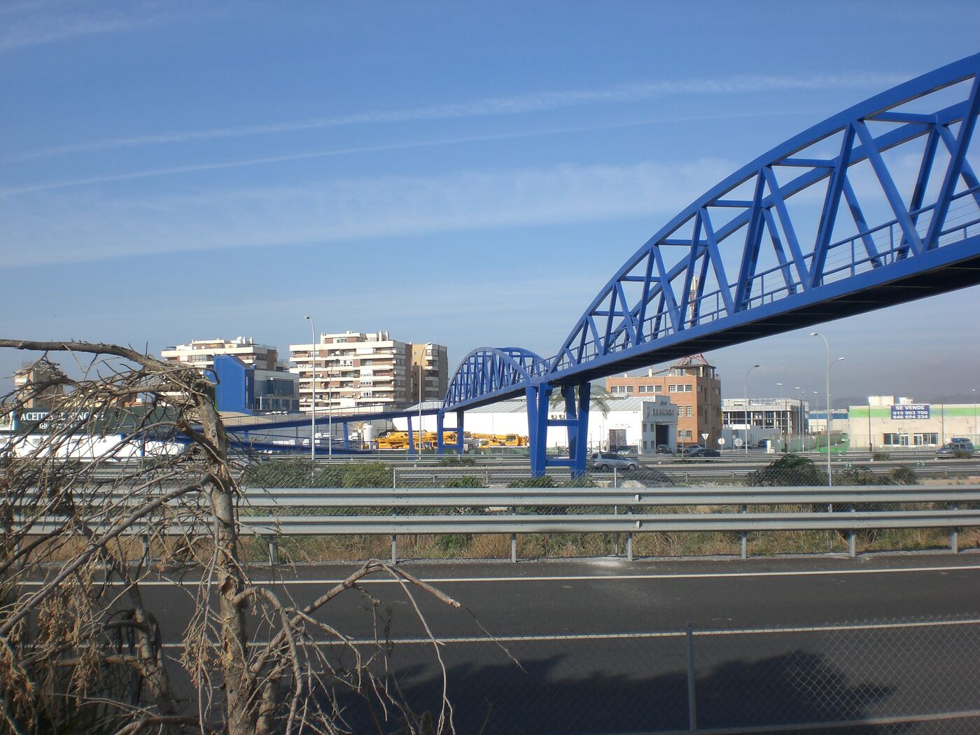 Pasarela peatonal metálica azul en arco sobre una autovía, con edificios al fondo